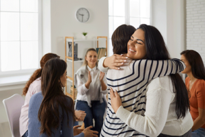 two people hug in a group therapy session in their drug detox treatment program while other members sit and talk with each other in the background