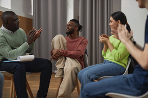 a man sits and shares his successes in his opioid and opiate addiction treatment program group therapy session while other group members sit in their chairs and clap