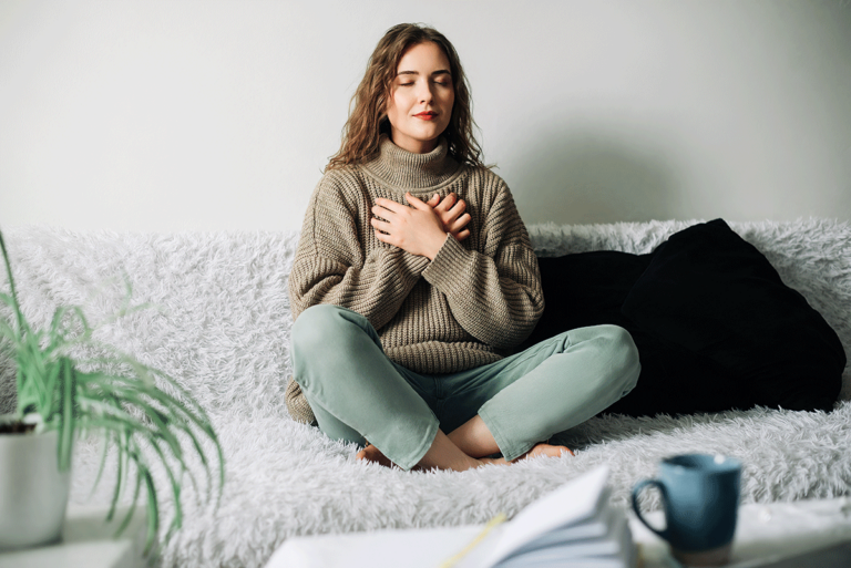 a young woman sits on her couch with her palms to her chest and meditating which is one of the coping skills for anxiety she learned